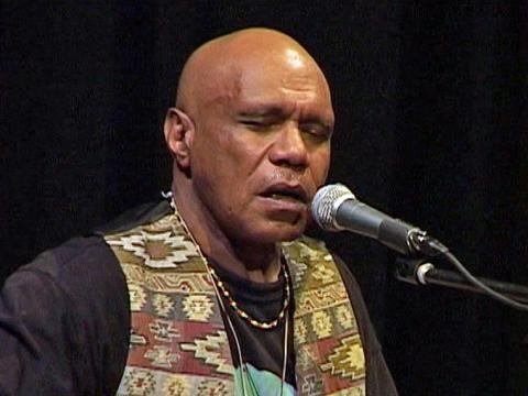 Head and shoulders shot of musician and songwriter Archie Roach seated in front of a microphone in front of a black backdrop. He is wearing a black shirt with a patterned vest.