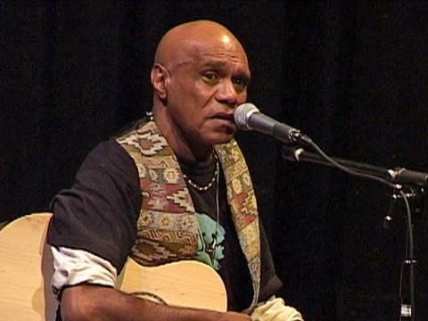Musician and songwriter Archie Roach seated in front of a microphone and holding an acoustic guitar in front of a black backdrop. He is wearing a black shirt with a patterned vest.