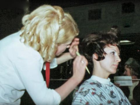 A hairdresser working on a woman's hair for a 1960s hairdressing competition. One of them has a white headpiece on the top of her head.