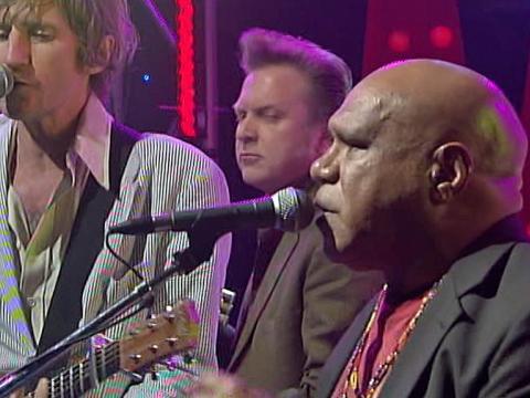 Indigenous musician Archie Roach pictured on stage with Tim Rogers, lead singer of the band You Am I and another musician in the background. Archie and Tim are both singing into microphones.