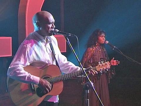 Indigenous musicians Archie Roach and Ruby Hunter pictured on stage performing. Archie is holding a guitar and Ruby is holding a tambourine and both are singing into microphones.