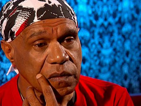 Close up of Indigenous singer-songwriter Archie Roach. He's wearing a headscarf and red t-shirt and has his hand on his chin.