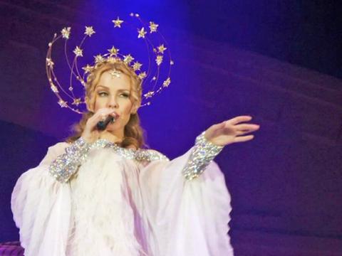 Kylie Minogue dressed in a white and silver gown with a silver, glittery head piece with stars on it. She's on stage singing into a microphone.