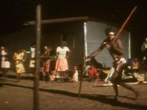 Man carrying a spear dancing at night with women in the background