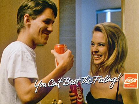 Kylie Minogue and a young man pictured from the chest up, drinking from cans of Coca-Cola and smiling at each other in a still from a Coca Cola cinema ad.