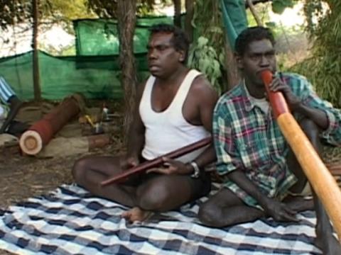 Two seated Aboriginal men, one playing clap sticks the other a didgeridoo