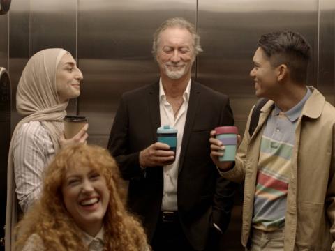 A female paralegal wearing a Hijab holds a coffee in an elevator. She is talking to two men also holding coffees and a laughing woman in a wheelchair