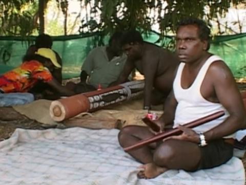 Aboriginal man seated playing clap sticks with others painting ceremonial objects