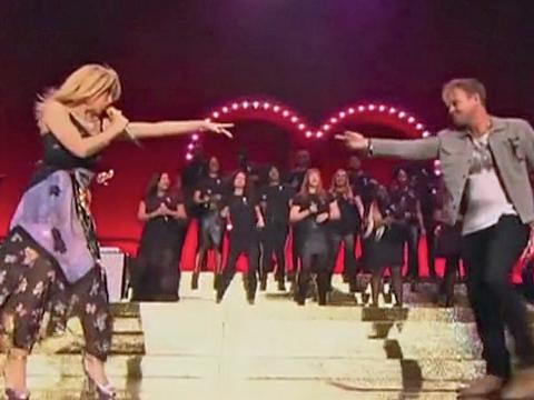 Kylie Minogue and Jason Donovan on a large stage together with arms outstretched toward eachother. In the background there is a heart-shape in pink lights and a choir dressed in black.
