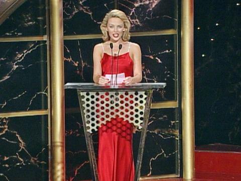 Kylie Minogue in a formal red gown standing behind a podium at an awards ceremony in 1999.