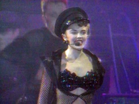 Kylie Minogue on stage singing wearing a sequined black bra and hat with a fishnet top.