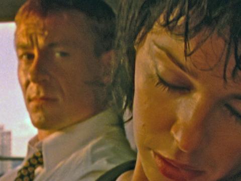 Close up of Kylie Minogue sleeping in a car with head tilted towards camera. Richard Roxburgh is sitting beside her looking at her. The still is from the short film Hayride to Hell. 