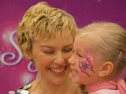Kylie Minogue smiles as she sits with a little girl next to her.