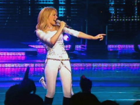 Kylie Minogue on stage holding a microphone to her mouth, dressed in a fitted white costume and she is pointing to one side of the stage