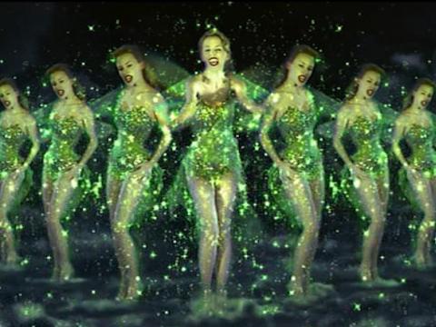 Eleven Kylie Minogues stand in a v formation wearing a costume that makes her look like a Green Fairy. She is singing.