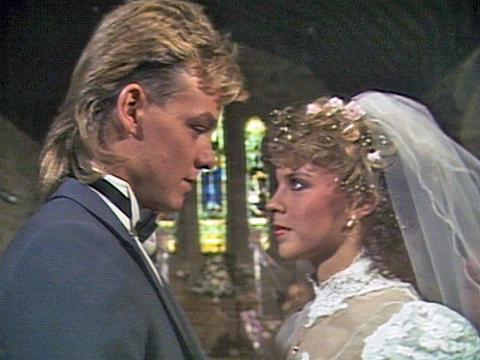 Jason Donovan and Kylie Minogue, pictured from shoulders up, appearing in character as Scott and Charlene in a scene from Neighbours where they get married. Jason is in a tuxedo and Charlene is wearing a lacy wedding gown and veil.