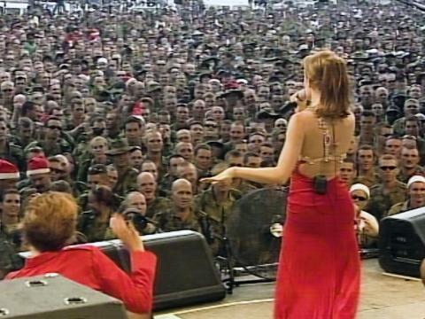 Kylie Minogue performing on a stage in a long, backless red dress, she has her back to camera facing out towards the large crowd.