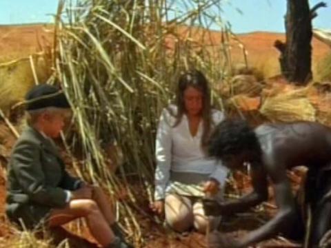 A young boy and teenage girl watch a young Aboriginal boy sucking water from the ground in the desert
