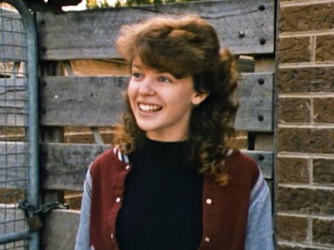 Kylie Minogue at age 17 pictured from the chest up, she is looking slightly away from the camera and smiling.