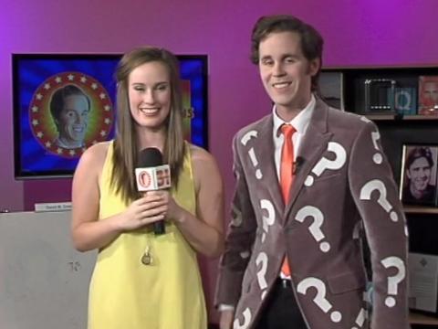 A young woman holding a microphone and man in a jacket with big question marks printed on it stand side by side smiling at the camera. They are hosting a TV game show.