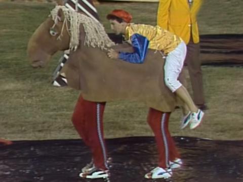 Two people in a horse costume and a person sitting on top of them dressed as a jockey