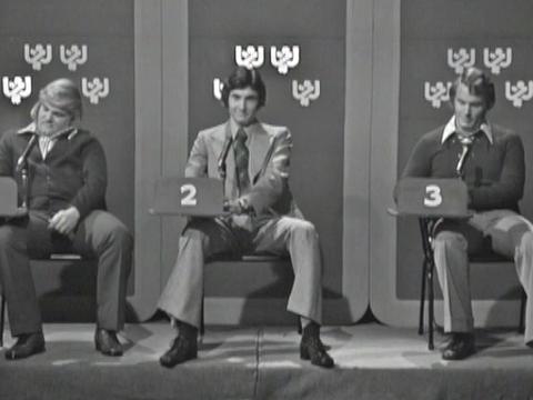 Three young men sitting on chairs in a row in a TV studio setting.