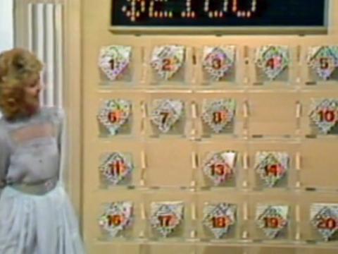 A woman standing next to a wall with rows of boxes on it in a game show setting.