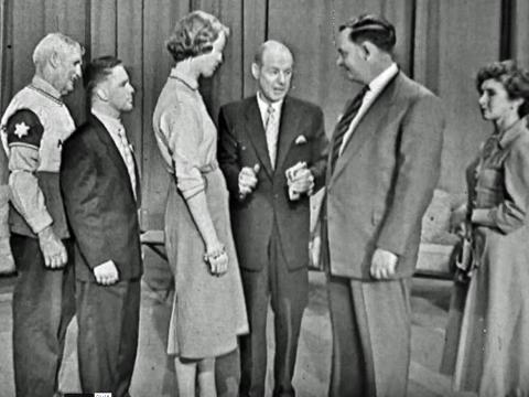 A row of men and women line up to play a tv game show with the host in the middle