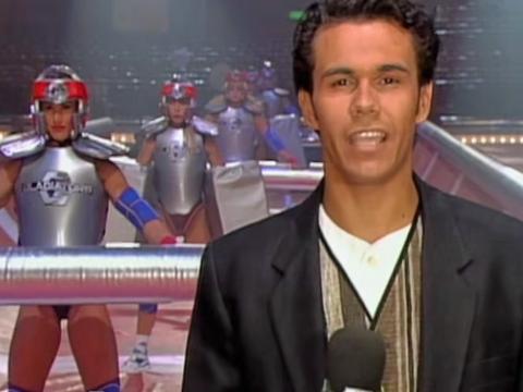 A young male TV presenter holding a microphone stands in front of a line of women dressed in battle gear.