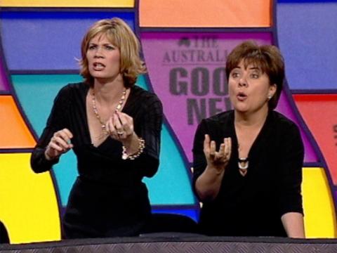 Two women dressed in black standing in front of a colourful wall in a TV studio. They are both holding out one hand and looking pleadingly at something off camera.