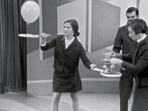 A woman holding tennis racquets in each hand. She bounces a balloon on one and another woman stacks cups and saucers on the other.