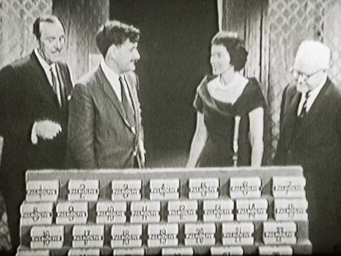 Three men and a woman on the set of a game show with rows of prize boxes in front of them.