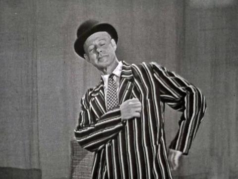A man in a bowler hat and striped jacket poses with his hand on his hip in a TV studio.