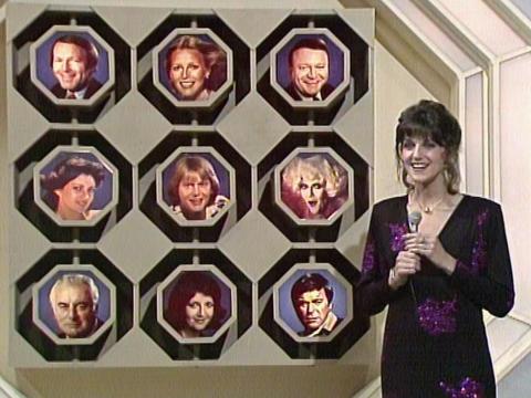 A woman game show co-host is holding a microphone and standing next to a board with the photos of nine celebrities on it.