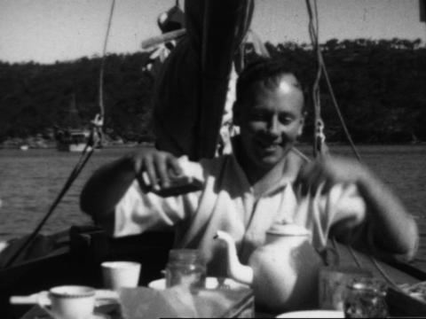Black and white still of a man sitting in a boat sailing on Sydney Harbour. He is taking tea from a china tea cup and teapot