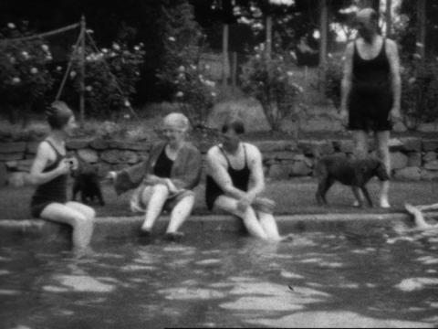 Black and white still people sitting around a swimming pool with their legs dipping in. Two dogs sit by the pool too.