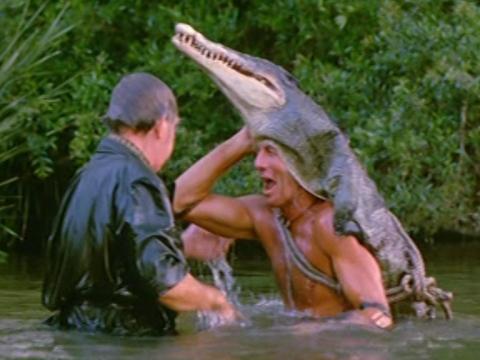 Paul Hogan as Mick 'Crocodile' Dundee laughs at Wally (John Meillon). Mick wears a crocodile skin, complete with head and jaws) on his head