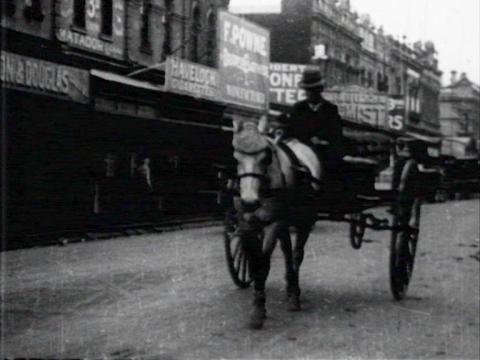 Black and white still of a man in a bowler hat driving a horse and cart down a street in Melbourne's Footscray in 1911