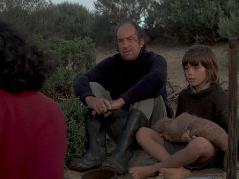 Peter Cummins (Hideaway Tom) and Greg Rowe (Storm Boy) are seated next to each other on the sand, talking with David Gulpilil (Fingerbone Bill) who his back to the camera.