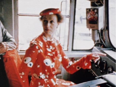 Princess Alexandra, wearing a bright-coloured, floral dress and hat, sitting in the driver's seat of a train and pointing to the controls.
