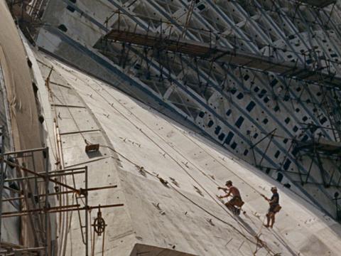 The Sydney Opera House's sail-like roof under construction with two workers scaling the massive roof.