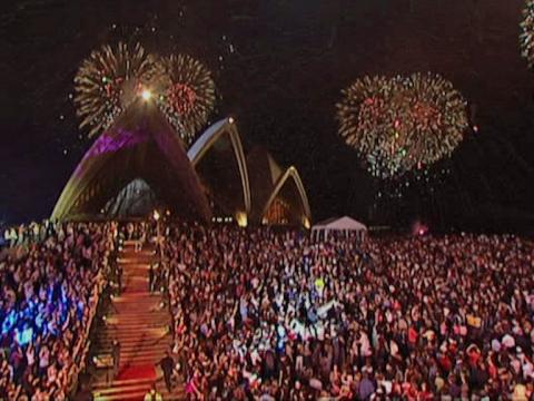 People cover the steps of the Sydney Opera House. The shell roofs peek over the top. There are fireworks in the night sky.