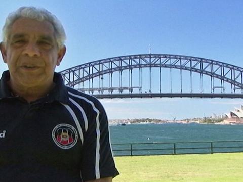A man is standing talking to camera. The Sydney Harbour Bridge and the Sydney Opera House are in the background.
