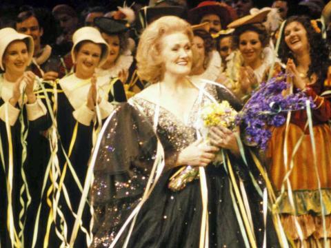 Dame Joan Sutherland is holding a bunch of flowers and is festooned in streamers. A crowd of people clap in the background.