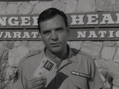Ed Deveraux in character speaking to camera holding some cards.