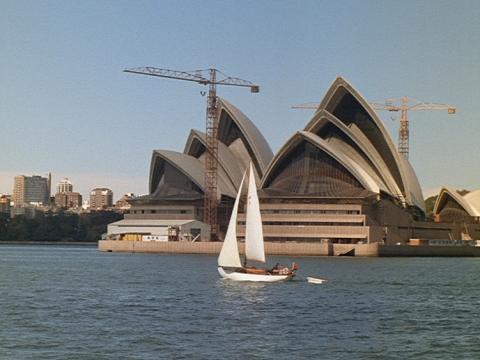 The Sydney Opera House with a sailing boat in the foreground. The House is still under construction and has two cranes alongside it.