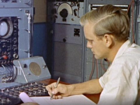 A technician takes notes in front of a console of switches and dials.