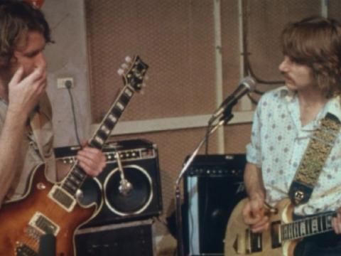 Two men holding electric guitars in a rehearsal room. Amplifiers in the background.