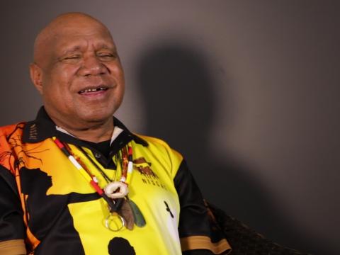 Archie Roach smiling, pictured from the waist up, seated and looking slightly away from camera.