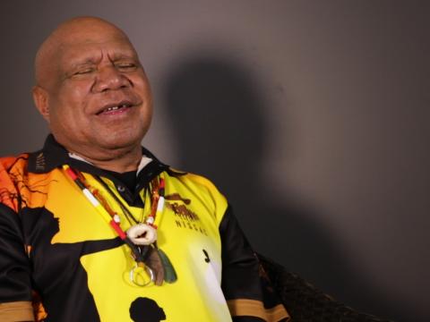 Archie Roach smiling, pictured from the waist up, seated and looking slightly away from camera.